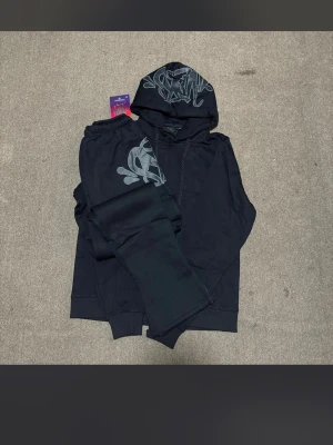 Svart hoodie (Syna world)  - Säljer en svart hoodie från (Syna world)med grått grafiskt tryck på huvan och bröstet. Hoodien har klassisk känguruficka, dragsko i huvan och ribbade muddar. Perfekt för dig som gillar streetwear och vill ha en stilren men ändå unik look.