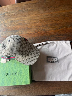 Gucci beige GG Supreme keps - Säljer en beige keps från Gucci i klassiskt GG Supreme-mönster. Kepsen har en böjd skärm och justerbar rem bak med grön-röd randig detalj. Materialet är canvas och kepsen kommer med originalförpackning och dustbag.