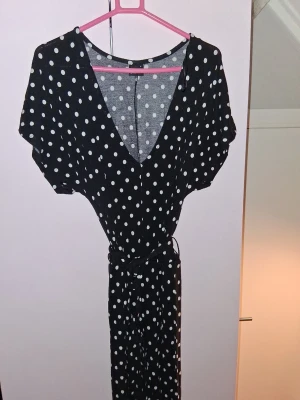 Svart prickig jumpsuit med knytband - Snygg svart jumpsuit med vita prickar och djup V-ringning. Korta ärmar och knytband i midjan för en smickrande siluett. Perfekt för dig som gillar en lekfull men stilren look. Tillverkad i ett mjukt och lätt tyg som känns skönt mot huden.