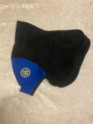 Svart och blå ansiktsmask - Säljer en svart ansiktsmask med blå detalj och en rund logotyp på sidan. Masken har en ergonomisk form som täcker näsa och mun, och är tillverkad i ett mjukt, flexibelt material. Perfekt för cykling eller utomhusaktiviteter när du vill skydda ansiktet mot vind och kyla. 95kr/st 