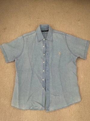 Blå linneskjorta från Ralph Lauren - Snygg ljusblå skjorta från Ralph Lauren i linne med korta ärmar och klassisk krage. Skjortan har knappar framtill och den ikoniska gula Polo-loggan broderad på bröstet. Perfekt för en avslappnad och stilren look.