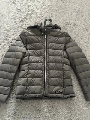 Grå jacka från Henri Lloyd - Snygg grå från Henri Lloyd med quiltad design och huva. Jackan har dragkedja framtill och en liten patch med brittisk flagga på nedre sidan. Perfekt för kalla vinterdagar och riktigt stilren look. Den har två fickor och den är storlek M men tycker den är väldigt liten och den sitter som en S
