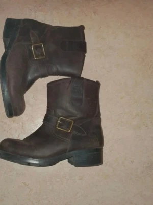 Bruna boots med spänne - Säljer ett par mörkbruna boots i läder med robust klack och dekorativa metallspännen på sidan. Klassisk design med rundad tå och dragflik upptill. Passar perfekt till jeans eller kjol för en cool look. Storlek 40
