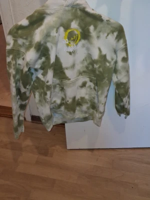 Grön tie-dye hoodie med smiley - Unik hoodie i grön och vit tie-dye med stor gul smiley på ryggen och texten 'NO RULES'. Mjuk bomullskänsla, huva och magficka. Perfekt för dig som gillar streetwear och vill sticka ut med en cool design.
