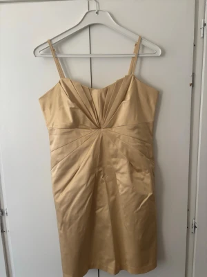 Champagnefärgad kort klänning i satin - Elegant kort klänning i champagnefärgad satin med smala axelband och veckade detaljer framtill som är snygg. Klänningen har en figurnära passform och en stilren look som passar perfekt till speciella tillfällen.