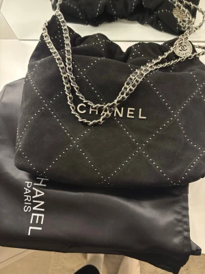 Svart Chanel handväska med kedja - Säljer en svart handväska från Chanel i mjukt mockaliknande material med silverfärgade nitar i rutmönster. Väskan har en kraftig silverkedja och en rund Chanel-tag. Loggan i metall framtill ger en lyxig känsla. Perfekt för dig som gillar exklusiva accessoarer.