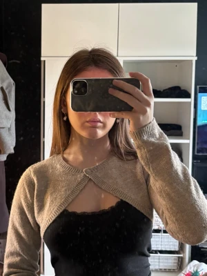 Beige kofta med knapp framtill - Kort beige bolero med lång ärm och en enkel knappstängning framtill. Tillverkad i mjukt stickat material som ger en avslappnad och trendig look. Perfekt att slänga över ett linne eller topp för extra stil.