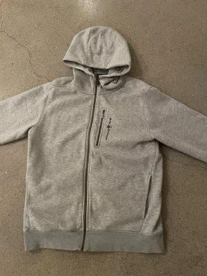 Grå hoodie från Sail Racing - Snygg ljusgrå hoodie från Sail Racing med dragkedja framtill, broderad logga på bröstet och en praktisk bröstficka med zip. Mjuk insida i fleece och två sidofickor. Perfekt för chill dagar eller när du vill ha en clean look.