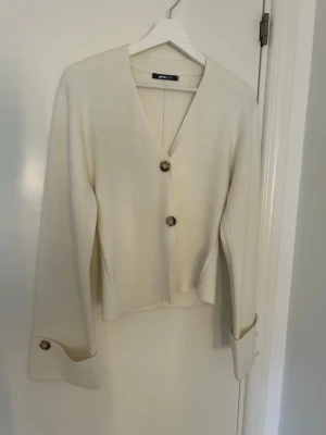 Krämvit kofta från Gina Tricot - Stilren krämvit kofta från Gina Tricot med v-ringning och stora, marmorerade knappar framtill och vid ärmsluten. Koftan har en rak passform och långa ärmar. Perfekt att slänga över en topp för en enkel men snygg look.