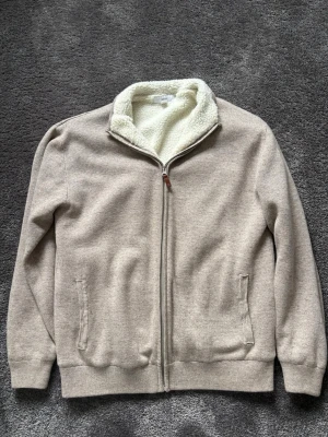 Beige ziptröja med teddyfoder - Säljer en beige ziptröja med mjukt vitt teddyfoder i kragen. Tröjan har hel dragkedja framtill, två fickor och ribbade muddar vid ärmslut och nederkant. Perfekt för dig som gillar en clean och mysig stil.