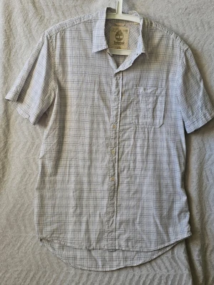 Vit rutig skjorta från Timberland - Klassisk vit kortärmad skjorta från Timberland med diskret rutigt mönster i blått och grått. Skjortan har en bröstficka, knappar framtill och en avslappnad regular fit. Perfekt till jeans eller shorts för en clean och enkel stil.