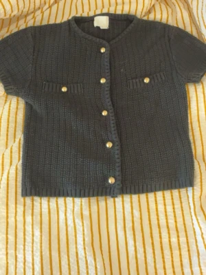 Svart kortärmad kofta från H&M - Kortärmad svart kofta från H&M med guldfärgade knappar framtill och dekorativa fickor på bröstet. Koftan har en rund halsringning och är stickad i ett grovt mönster. Perfekt för att lyfta en enkel outfit.