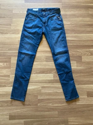 Blå slim fit jeans från Replay - Säljer ett par blå slim fit jeans från Replay med klassisk femficksdesign och dragkedja fram. Jeansen har raka ben och är tillverkade i mjuk denim med snygga sömmdetaljer på bakfickorna. Perfekta för en avslappnad och trendig look. 
