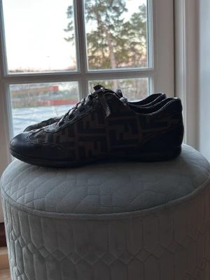 Bruna Fendi sneakers med FF-mönster - Säljer ett par bruna sneakers från Fendi med det klassiska FF-mönstret i tyg och detaljer i läder. Skorna har låga skaft, snörning och en stilren design som sticker ut. Perfekta för dig som gillar lyxiga och unika sneakers. Nypris 4000.