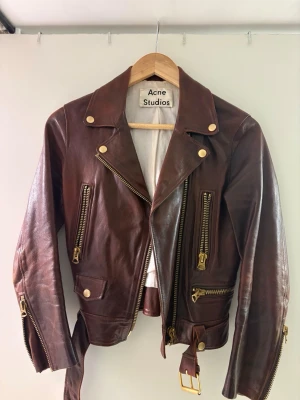 Brun skinnjacka från Acne Studios - Snygg brun skinnjacka från Acne Studios med guldiga dragkedjor och knappar. Klassisk bikerstil med bred krage, bälte i midjan och flera fickor framtill. Jackan har en cool vintagekänsla och detaljer som ger extra edge till din outfit. Kostade 8000kr ny. 
