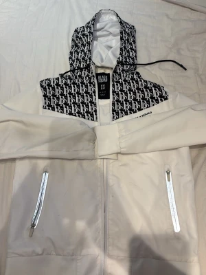 Vit vindjacka från Black Squad - Snygg vit vindjacka från Black Squad med svartvit huva och axelparti i BS-logga. Jackan har dragkedja framtill, två fickor med vita dragkedjor och elastiska muddar vid ärmarna. Perfekt för en streetwear-look och enkel att matcha.