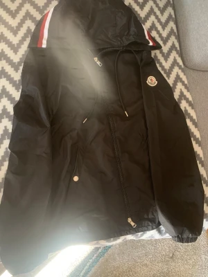 Moncler vindjacka äkta  - Svart vindjacka från Moncler med huva och dragkedja framtill. Jackan har vita och röda ränder på axlarna, justerbar dragsko och Moncler-logga på bröstet. Tillverkad i lätt, vattenavvisande material som passar perfekt för blåsiga dagar.