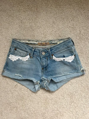 Blå jeansshorts med spetsdetaljer - Säljer ett par ljusblå jeansshorts med vita spetsdetaljer vid fickorna. Midjemåttet rakt över :  34 cm och längden på shortsen     : 23 cm. Kan ej visa på eftersom dom inte passar mig. Hör av er vid några funderingar!