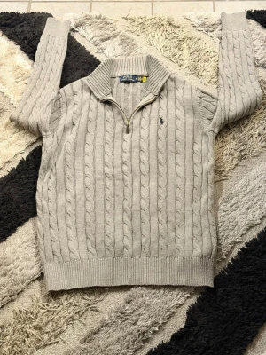 Grå kabelstickad tröja Polo Ralph Lauren - Snygg grå kabelstickad tröja från Polo Ralph Lauren med halv dragkedja och klassisk blå logga broderad på bröstet. Tröjan har ribbade muddar och krage, perfekt för lager på lager. Materialet känns mjukt och håller värmen.