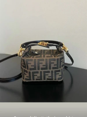 Fendi brun FF-mönstrad handväska - Säljer en brun handväska från Fendi med det klassiska FF-monogrammet i mörkbrunt och svart. Väskan har guldfärgade metalldetaljer, avtagbar axelrem och ett kort handtag i läder. Perfekt för dig som gillar lyxiga accessoarer och vill sticka ut.