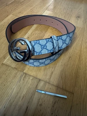 G  bälte  - Snyggt bälte med klassiskt mönster i blått och vitt. Bältet har ett stort, rundat spänne i silverfärg. Insidan är beige och materialet är skinn. Perfekt accessoar för att lyfta din outfit.