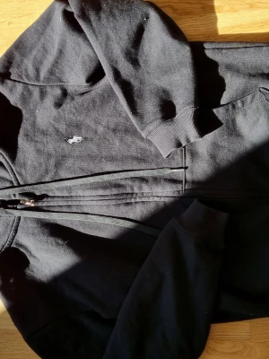 Svart zip-hoodie från Polo Ralph Lauren - Klassisk svart hoodie med dragkedja från Polo Ralph Lauren. Hoodien har huva, ribbade muddar och fickor framtill. Den ikoniska broderade loggan i vitt syns på bröstet. Perfekt för en avslappnad och sportig stil.
