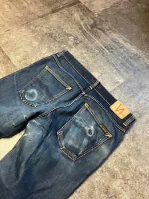 Blå jeans från Nudie Jeans - Säljer ett par klassiska blå jeans från Nudie Jeans med snygg slitning och kontrastsömmar. Modellen har raka ben, fem fickor och sköna fades. Sitter mindre så hör av er vid intresse kring måtten!