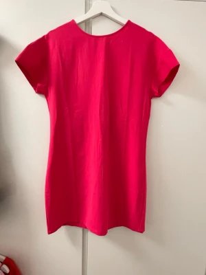 Skrikrosa kortklänning med djup v-ringning i ryggen - Säljer en snygg rosa kortklänning med djup v-ringning i ryggen och korta ärmar. Klänningen har en enkel, rak passform och är tillverkad i ett mjukt bomullstyg. Perfekt för dig som vill ha en stilren men ändå iögonfallande look.