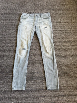 Jeans - Säljer ett par ljusgrå skinny jeans med slitningar och hål på benen för en trendig, avslappnad look. Jeansen har klassisk femficksdesign och knappgylf. Perfekta för dig som gillar streetstyle och vill ha ett par riktigt snygga jeans med attityd.