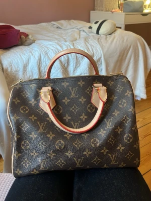 Louis Vuitton Speedy Monogram väska - Klassisk Louis Vuitton Speedy handväska i brunt monogramcanvas med beige och röda läderdetaljer. Väskan har två handtag, guldfärgade metalldetaljer och dragkedja upptill. Ikoniskt LV-mönster över hela väskan. Perfekt för dig som gillar lyxig och tidlös design.