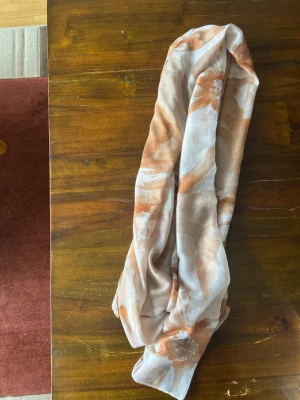Beige och orange mönstrad sjal - Säljer en tunn och mjuk sjal i beige och orange med abstrakt mönster. Sjalen är rektangulär och passar perfekt att svepa runt halsen för en snygg touch till din outfit. Materialet känns lätt och luftigt, vilket gör den enkel att styla på olika sätt.
