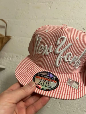 Randig New York keps från CityHunter - Två snygga snapback-kepsar från CityHunter med broderad 'New York'-text framtill. En keps är röd- och vitrandig, den andra grön- och vitrandig. Klassisk flat brim och cool streetwear-vibe. Tillverkad i bomullsmix med tydliga etiketter och stickers kvar. 135kr/st 