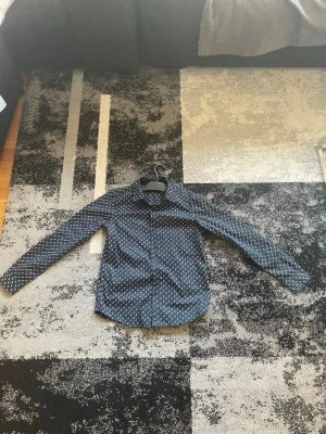 Blå mönstrad skjorta med knappar - Snygg blå skjorta med vitt prickigt mönster och klassisk krage. Skjortan har långa ärmar och knäppning framtill. Perfekt för dig som vill ha en stilren men ändå unik look.