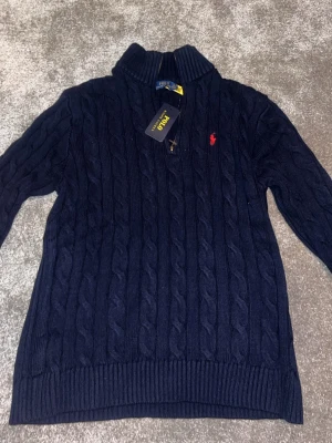 Mörkblå kabelstickad tröja Polo Ralph Lauren - Snygg mörkblå kabelstickad tröja från Polo Ralph Lauren med halv dragkedja och hög krage. Tröjan har det klassiska röda Polo-logot på bröstet och ribbade muddar vid ärmslut och nederkant. Perfekt för dig som gillar stilren och tidlös design. (Rep)