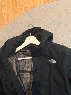 Svart regnjacka från The North Face - Snygg svart regnjacka från The North Face med huva och meshfoder. Jackan har dragkedja, tryckknappar och två fickor framtill. Loggan är tryckt på bröstet och ärmen. Perfekt för regniga dagar och outdoor-äventyr.