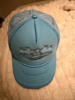 Blå truckerkeps med stjärnor - Snygg ljusblå truckerkeps med mesh på sidorna och broderade stjärnor framtill. Kepsen har böjd skärm och justerbar baksida för perfekt passform. Materialet är en mix av tyg och mesh som ger en luftig känsla.