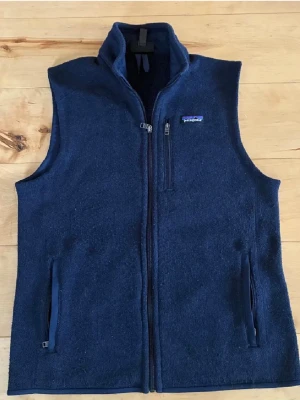 Mörkblå dunväst från Patagonia - Mörkblå dunväst från Patagonia med hel dragkedja, hög krage och tre fickor med dragkedja. Västen har en klassisk, sportig look och är tillverkad i ett mjukt, värmande material. Perfekt för lager-på-lager-stil under kyliga dagar.