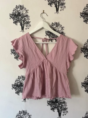 Ljusrosa blus med volangärm och knyt - Supersöt ljusrosa blus med v-ringning både fram och bak, volangärmar och knytband i nacken. Tillverkad i ett tunt, skrynkligt bomullsliknande material som ger en luftig känsla. Perfekt för varma dagar och ger en romantisk vibe.