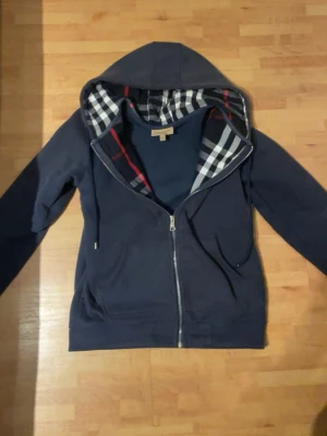 Marinblå hoodie från Burberry - Snygg marinblå hoodie från Burberry med dragkedja och klassiskt rutigt foder i huvan. Hoodien har fickor framtill, justerbar dragsko i huvan och ribbade muddar. Perfekt för dig som vill ha en stilren och bekväm look med en touch av lyx. Begagnad men är i mycket bra skick!