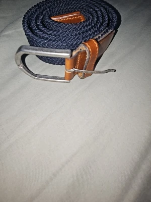 Flätat bälte - Snyggt mörkblått flätat bälte med detaljer i brunt skinn och silverfärgat spänne. Perfekt accessoar för att lyfta din outfit och funkar till både jeans och chinos. Klassisk design med modern twist.