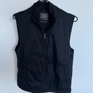 Svart väst från Berkeley - Stilren svart dunväst från Berkeley med hög krage och dragkedja framtill. Västen har två sidofickor och en enkel, klassisk design som passar till många olika outfits. Perfekt för lager-på-lager under kyligare dagar.