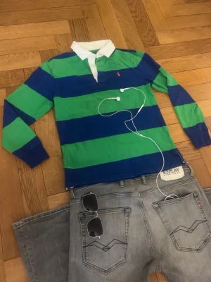 Randig rugbytröja från Ralph Lauren | long sleeve | barn | - Säljer en klassisk långärmad rugbytröja från Ralph Lauren med breda gröna och blå ränder, vit krage och knappslå. Tröjan har en liten röd logga broderad på bröstet och är gjord i mjuk bomul. Perfekt för en sportig och avslappnad stil.