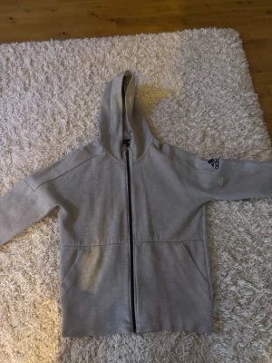 Grå zip hoodie från Adidas - Säljer en grå hoodie från Adidas med dragkedja framtill och två stora fickor. Tröjan har huva och Adidas-logga på ena ärmen. Tillverkad i mjukt sweatshirtmaterial, perfekt för en avslappnad stil.