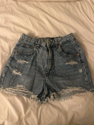 Högmidjade slitna jeansshorts - Säljer ett par högmidjade blå jeansshorts med slitna detaljer och fransig kant. Klassisk femficksmodell med dragkedja och knapp framtill. Perfekta för sommaren och ger en avslappnad vibe.