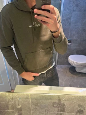 Olivgrön hoodie Ralph Lauren - Snygg olivgrön hoodie från Ralph Lauren med broderad vit text på bröstet. Klassisk känguruficka framtill, justerbar huva med snören och ribbade muddar. Perfekt nu till våren. Jag på bilden är 188cm och bör storlek S vilket är ganska litet på mig. Hör av er vid frågor:)