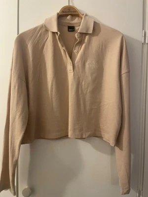 Helt ny Gina Tricot Beige croppad långärmad polotopp - Oanvänd sportig topp med knappar till halsen. Toppen är beige och långärmad och har en krage. Den har en kort, boxig passform. Tvättad men inte använd.