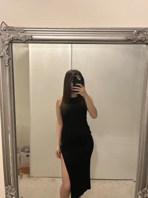 Svart långklänning med slits - Säljer en svart långklänning i figurnära modell med hög slits på ena sidan. Klänningen är ärmlös och har en enkel, stilren design som passar perfekt till dig som gillar minimalistiskt mode.