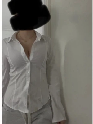 Vit figurnära skjorta med knappar - Klassisk vit skjorta i figurnära modell med lång ärm och knäppning framtill. Skjortan har en djup v-ringning och krage, perfekt för en stilren look. Tillverkad i ett tunt bomullstyg som känns bekvämt och följsamt.