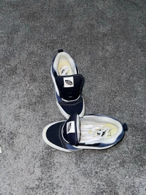 Marinblå och vita Vans sneakers - Säljer ett par klassiska Vans sneakers i marinblått och vitt. Skorna har vit sula, vita skosnören och den ikoniska Vans-loggan på plösen. Ovandelen är i canvas och mocka, vilket ger en snygg kontrast. Perfekta för en avslappnad streetwear-look.