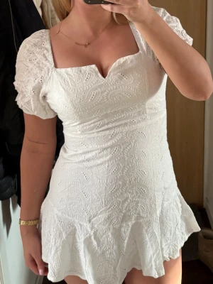 Vit kort klänning med volang - Söt vit kortklänning med broderade detaljer och volangkant. Klänningen har puffiga kortärmar och en snygg v-ringad urringning. Perfekt för sommarens alla tillfällen och ger en romantisk vibe.